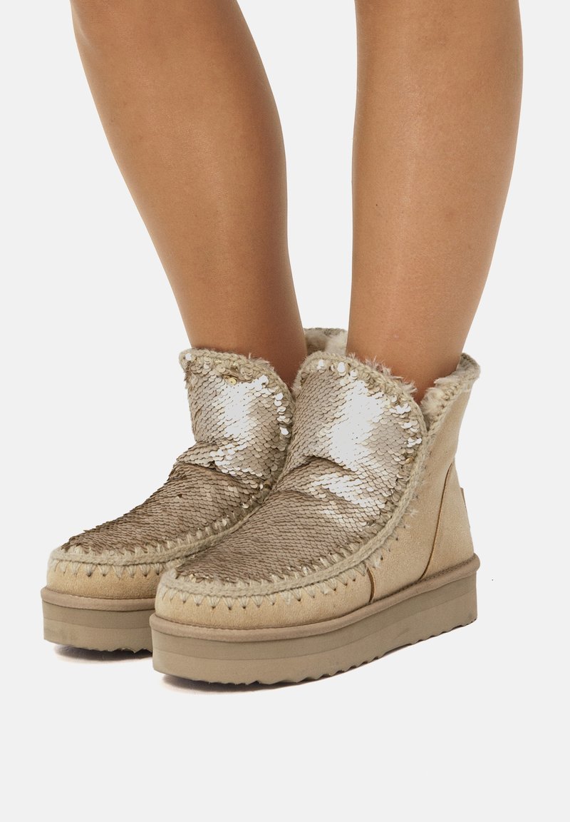 Stivaletti alla caviglia dorati con paillettes, foderati in pelliccia sintetica, base in suede beige e cuciture decorative. Suola piatta con grip testurizzato.