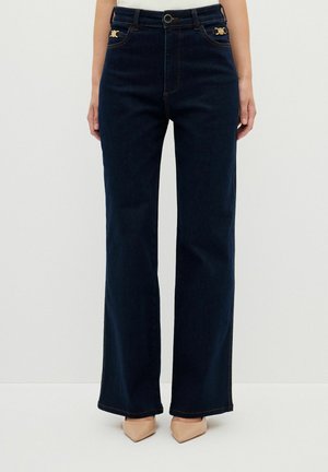 Kvinde iført mørkeblå højtaljede jeans med vide ben og gyldne knapdetaljer nær lommerne samt beige spidse sko.