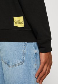 Sudadera negra con un parche del logo amarillo de "Los Simpson" en el dobladillo, combinada con jeans claros desgastados. Textura de algodón suave visible.