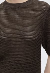 Gros plan d'une personne portant un pull en maille marron foncé à manches courtes avec un motif texturé et à tissage ouvert.