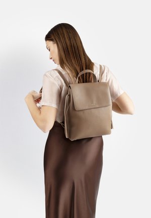 Donna che regola gli spallacci di uno zaino in pelle taupe, indossa una camicetta chiara e una gonna marrone scuro, voltata di spalle su sfondo bianco uniforme.