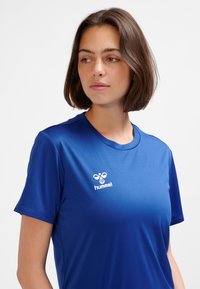 Koszulka sportowa z krótkim rękawem w jaskrawym niebieskim kolorze, wykonana z gładkiego materiału, z białym logo po lewej stronie klatki piersiowej.