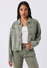 Groene denimjas met een textuurpatroon, knoopsluiting en rauwe zoom. Draagt over een witte top, gecombineerd met bijpassende groene broek.