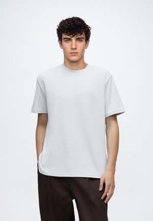 Junger Mann mit lockigem dunklem Haar, der ein schlichtes weißes kurzärmeliges T-Shirt und dunkle Hosen trägt, vor einem hellen Hintergrund stehend.
