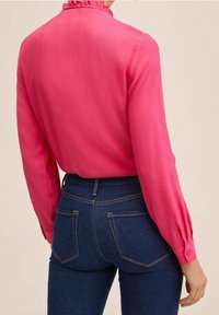 Blusa rosa a maniche lunghe con colletto arricciato, tessuto morbido e polsini con bottoni, abbinata a jeans skinny blu scuro con tasche posteriori.