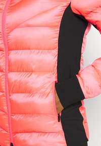 Zbliżenie na osobę noszącą pikowaną kurtkę w odcieniu koralowo-różowym i czarnym, z ręką w zapinanej na zamek kieszeni bocznej.