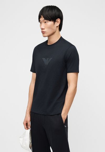 Νεαρός άντρας που στέκεται, φοράει μαύρο μπλουζάκι με διακριτικό λογότυπο στο στήθος, μαύρο παντελόνι, κρατώντας ένα λευκό καπέλο στο ένα χέρι.