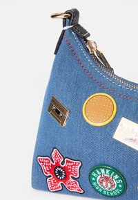 Denimväska med triangulär form, prydd med olika färgglada lappar: en kassettband, ett våffla, en blomma och en skollogga. Guldzipperdetalj.