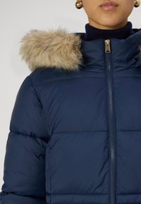 Jūras spēku puffer jaka ar mākslīgās kažokādas kapuci, ar priekšējo rāvējslēdzēju, quilted tekstūru un ribotu apkakles detaļu.