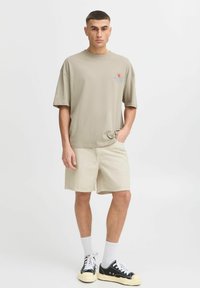 Beige Baumwoll-T-Shirt mit kurzen Ärmeln und grafischem Design, kombiniert mit hellbeigen Shorts. Schwarze Sneakers mit weißen Schnürsenkeln und hellen Socken.