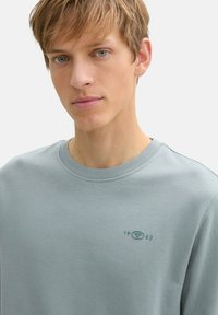 Hellblaues Sweatshirt mit Rundhalsausschnitt, ausgestattet mit einem geprägten Logo und dem Jahr "1962" auf der Brust. Glatte Textur und lässige Passform.