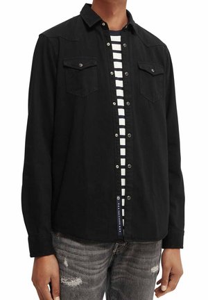 Homme portant une chemise noire à boutons par-dessus un t-shirt rayé noir et blanc et un jean gris déchiré.