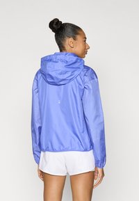 Ελαφρύ μπλε μπουφάν με κουκούλα, με μανίκια σε σχέδιο από δίχτυ. Το ύφασμα έχει ομαλή υφή και το σχέδιο περιλαμβάνει ανακλαστική λεπτομέρεια στην πλάτη.