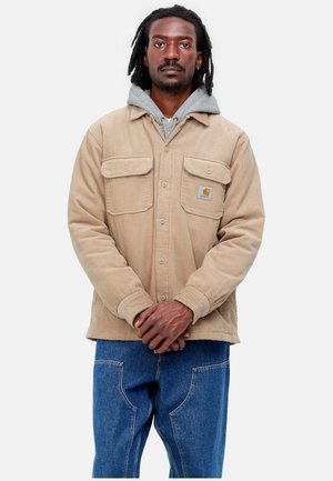 Chaqueta de pana beige con dos bolsillos en el pecho, cierre de botones al frente y una sudadera con capucha gris por debajo. Los pantalones son de mezclilla azul.