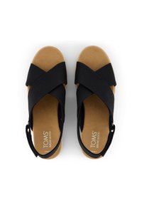 Un par de sandalias de mujer en color marrón claro con tiras negras entrelazadas y correas en el tobillo, con la marca TOMS visible en las plantillas, visto desde arriba.