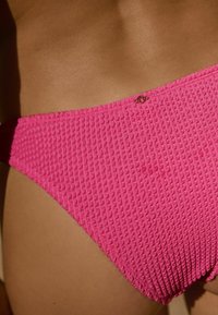 Rozā teksturētas bikini apakšdaļa ar viļņotu rakstu, kas ir ar nelielu metāla akcentu. Dizainā ir plaša josta atbalsta nodrošināšanai.