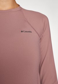 Ilgųjų rankovių sportinė marškinėliai ramios mauve spalvos su lygia tekstūra. Ant krūtinės yra subtilus prekių ženklų logotipas ir kontrastingi siūlų detalės.