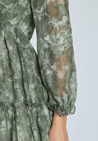 Vestido verde con estampado floral hecho de tela transparente, con puños de mangas con volantes y tela fruncida en la cintura, que presenta intrincados detalles de bordado.