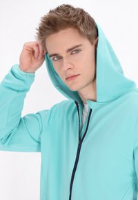 Felpa blu chiaro con una texture morbida, chiusura con zip frontale e un ampio cappuccio. Il modello sta sistemando il cappuccio, mettendo in evidenza il suo design.