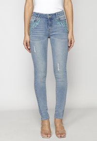 Jeans ajustados de mezclilla azul claro con cuentas decorativas en los bolsillos frontales, detalles desgastados en las rodillas y un corte ceñido.