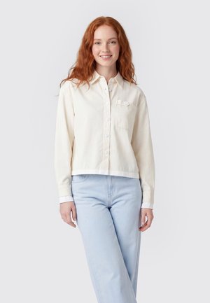 Lächelnde rothaarige Frau mit weißem Hemd und hellblauen Jeans stehend vor einem schlichten hellen Hintergrund.