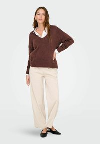 Donna che indossa un maglione marrone a scollo a V sopra una camicia bianca con colletto, pantaloni beige a gamba larga e scarpe nere a punta, in piedi con una mano sulla vita.