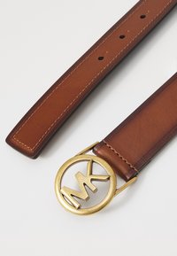 Cintura in pelle marrone con fibbia circolare dorata che presenta il logo "MK". Texture liscia, cuciture visibili e più fori per la regolazione.