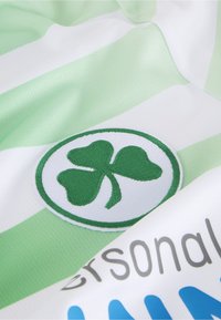 Textil de rayas verdes y blancas con un logotipo de trébol verde bordado sobre un fondo blanco circular; tejido suave con una textura lisa.