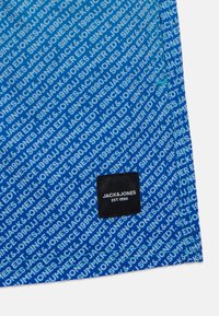 Zila auduma, kurā atkārtojas balts teksts "JACK & JONES" un "SINCE 1990." Apakšējā stūrī ir melna birka ar balto tekstu.