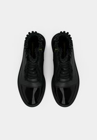 Schwarze Stiefel bis zum Knöchel mit einer glänzenden Lackspitze, einem strukturierten Leder-Obermaterial, gerüschten Knöchelverzierungen und einem geprägten Logo auf der Innensohle.