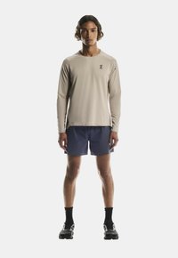 Beige langärmliges Hemd mit einem Logo, kombiniert mit marineblauen Shorts. Das Modell trägt schwarze Sportschuhe und Socken. Einfaches, funktionales Design.