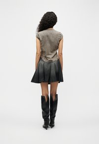 Γυναίκα με κατσαρά μαλλιά, που φοράει γκρι εφαρμοστό αμάνικο τοπ, μαύρη πλισέ μίνι φούστα και μαύρες μπότες με τακούνι μέχρι το γόνατο, στραμμένη προς τα πίσω.