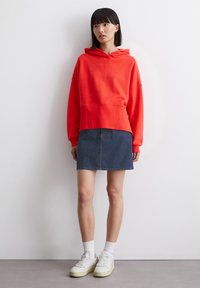 Rode hooded sweatshirt met een relaxed fit, gecombineerd met een donkere denim rok. Het model draagt witte sneakers en crewsokken.