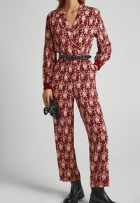 Combinaison rouge à motifs avec manches longues, taille cintrée et ceinture noire ; présente un design géométrique et des poignets rouges unis. Portée avec des bottines noires.