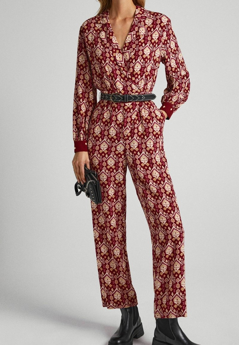 Combinaison rouge à motifs avec manches longues, taille cintrée et ceinture noire ; présente un design géométrique et des poignets rouges unis. Portée avec des bottines noires.