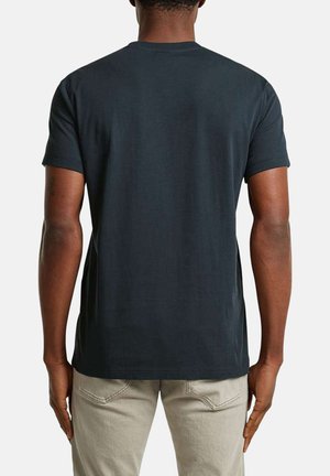 Homme portant un t-shirt à manches courtes gris foncé uni et un pantalon beige, vu de dos, debout devant un fond blanc.