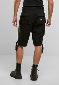 Mann som har på seg svarte cargo shorts med baklommer og svarte støvler, stående på et lys grått gulv mot en ensfarget bakgrunn.