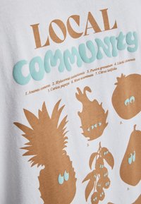 Λευκό T-shirt με παιχνιδιάρικα καφέ και γαλάζια φρουτώδη σχέδια, όπως ανανά και διάφορα φρούτα, με την επιγραφή "ΤΟΠΙΚΗ ΚΟΙΝΟΤΗΤΑ."