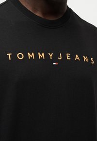 Μαύρο βαμβακερό t-shirt με χρυσή κεντημένη επιγραφή "TOMMYJEANS" και μικρό λογότυπο σημαίας σε κόκκινο, λευκό και μπλε στο ύφασμα.