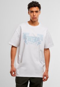 Joven con pelo rizado que lleva una camiseta blanca de gran tamaño con texto decorativo azul claro y pantalones negros, frente a un fondo liso.