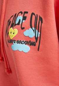 Sudadera color coral con un corazón sonriente y nubes azules, con el texto "PAZ SIEMPRE A RITMO" en negro. Tejido suave, diseño tipo pullover.