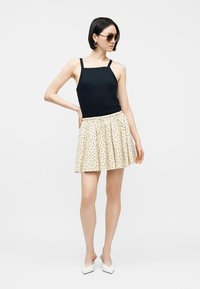 Γυναίκα που φοράει μαύρο αμάνικο τοπ με τετράγωνο ντεκολτέ, μπεζ φούστα με πουά, λευκά μυτερά παπούτσια, γυαλιά ηλίου και κρίκους στα αυτιά, ποζάροντας μπροστά σε λευκό φόντο.