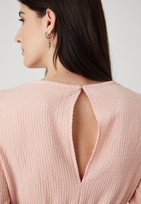 Rosa teksturert topp med en ryggåpning sikret med en liten knapp, korte ermer og et bølget stoffdesign.