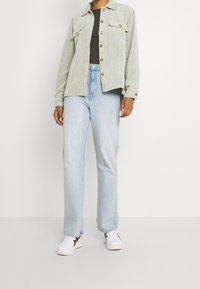 Ljust grön kordflöjelskjorta i jackmodell med två bröstfickor, i kombination med ljusblå raka jeans och vita sneakers med svarta accenter.