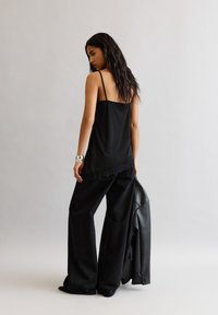 Hosszú, sötét hajú nő fekete csipkével díszített topban és széles fekete nadrágban, egy fekete bőrdzsekit tart a kezében, egy sima világos háttér előtt.