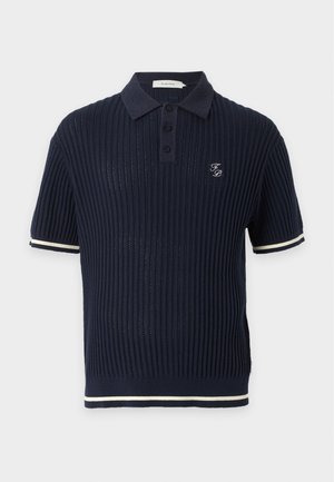 Homme portant un polo noir côtelé à manches courtes, un jean bleu foncé ample et des baskets blanches, debout avec une main dans la poche.