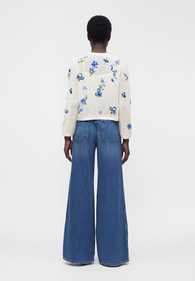 Person som bär en vit stickad tröja med blå blommönster och vida blå jeans, står med ryggen mot en vit bakgrund.