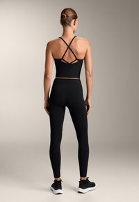 Čierny športový crop top s krížovými popruhmi, v kombinácii s čiernymi legínami. Model nosí čierne tenisky, ktoré ukazujú hladký, priliehavý dizajn.
