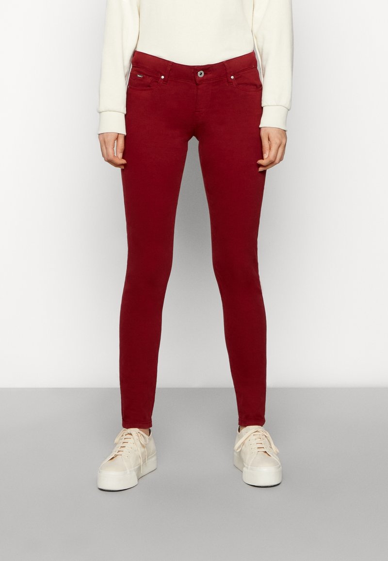 Slim-fit tmavě červené kalhoty, které jsou kombinovány s bílým topem s dlouhým rukávem a světlým platformovým teniskam na jednobarevném pozadí.