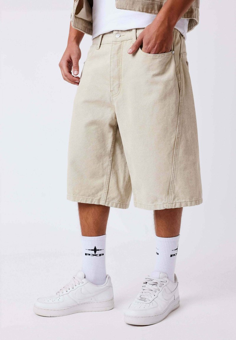 Homme portant un short beige jusqu'aux genoux, des chaussettes blanches montantes avec un logo d'avion noir, et des baskets blanches, debout avec une main dans la poche.